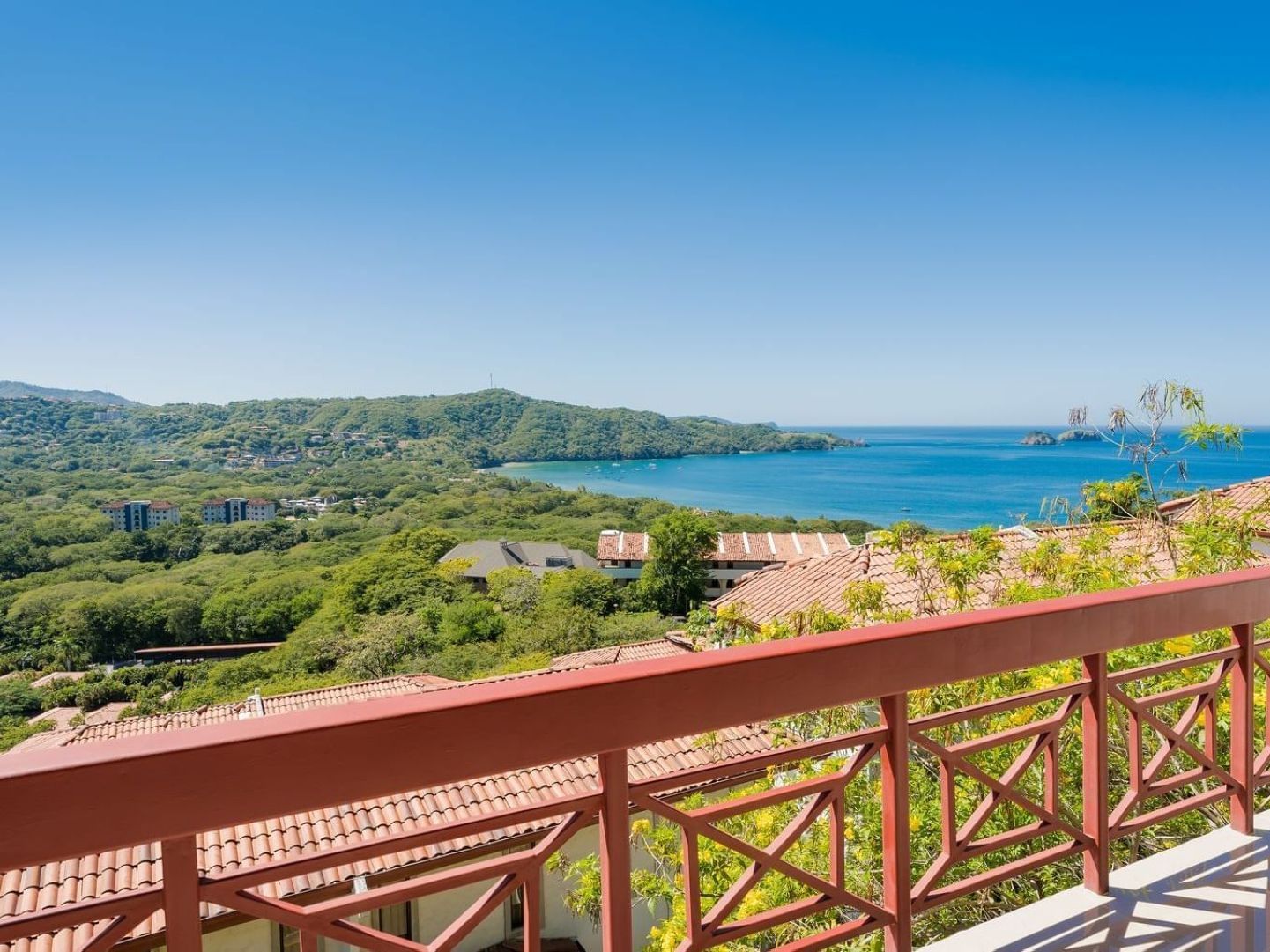 Hillside resort property overlooking turquoise bay with red-roofed buildings, dense green vegetation, and forested coastline under clear sky