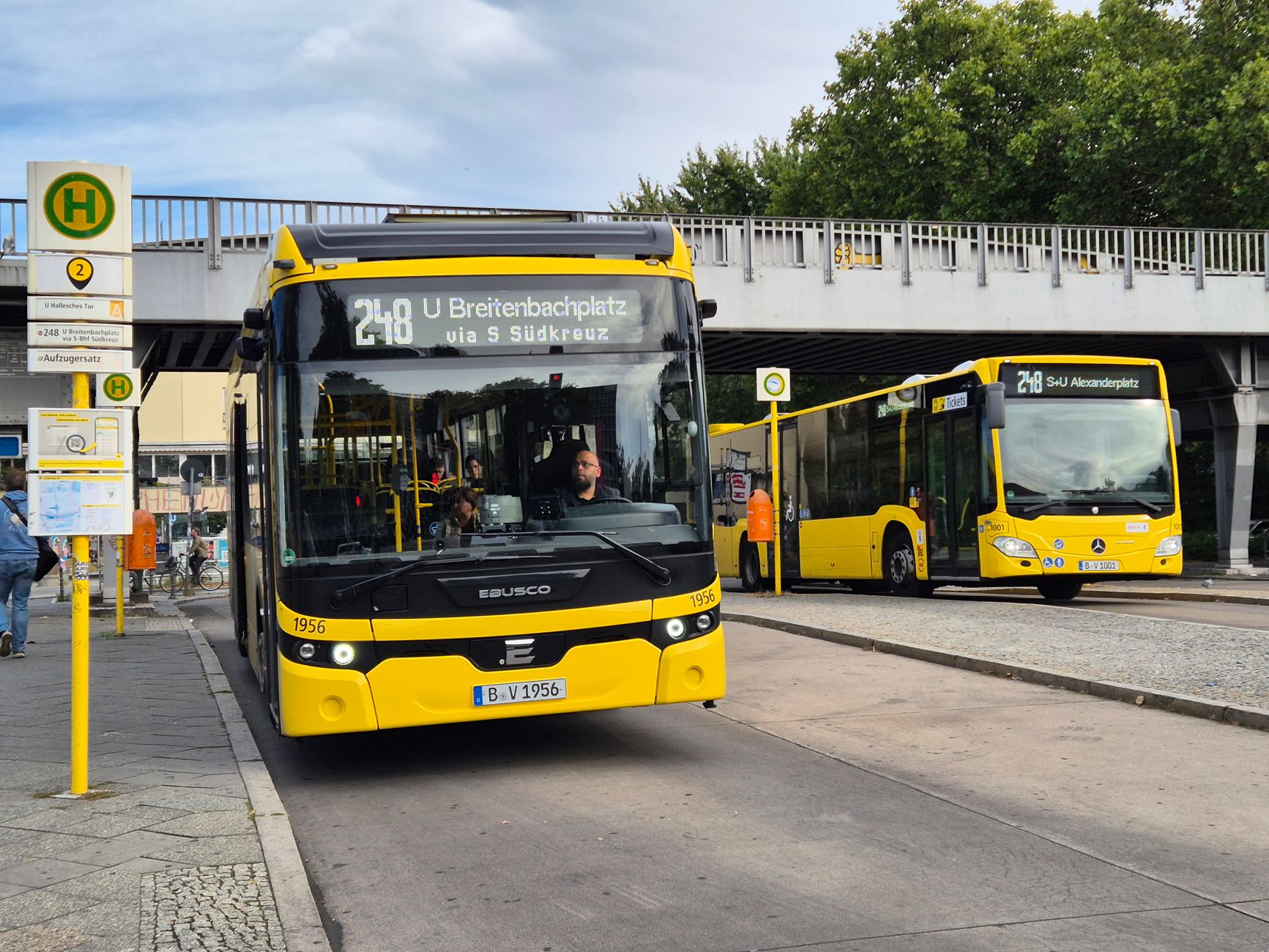 Gelbe Busse halten an einer Bushaltestelle unter einem Bahnviadukt in einer deutschen Stadt.