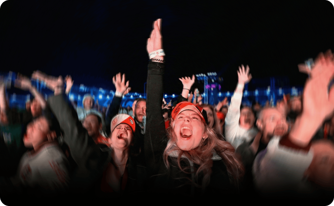 Excited fans wearing red caps cheering with raised arms at a nighttime outdoor event with blue stage lighting