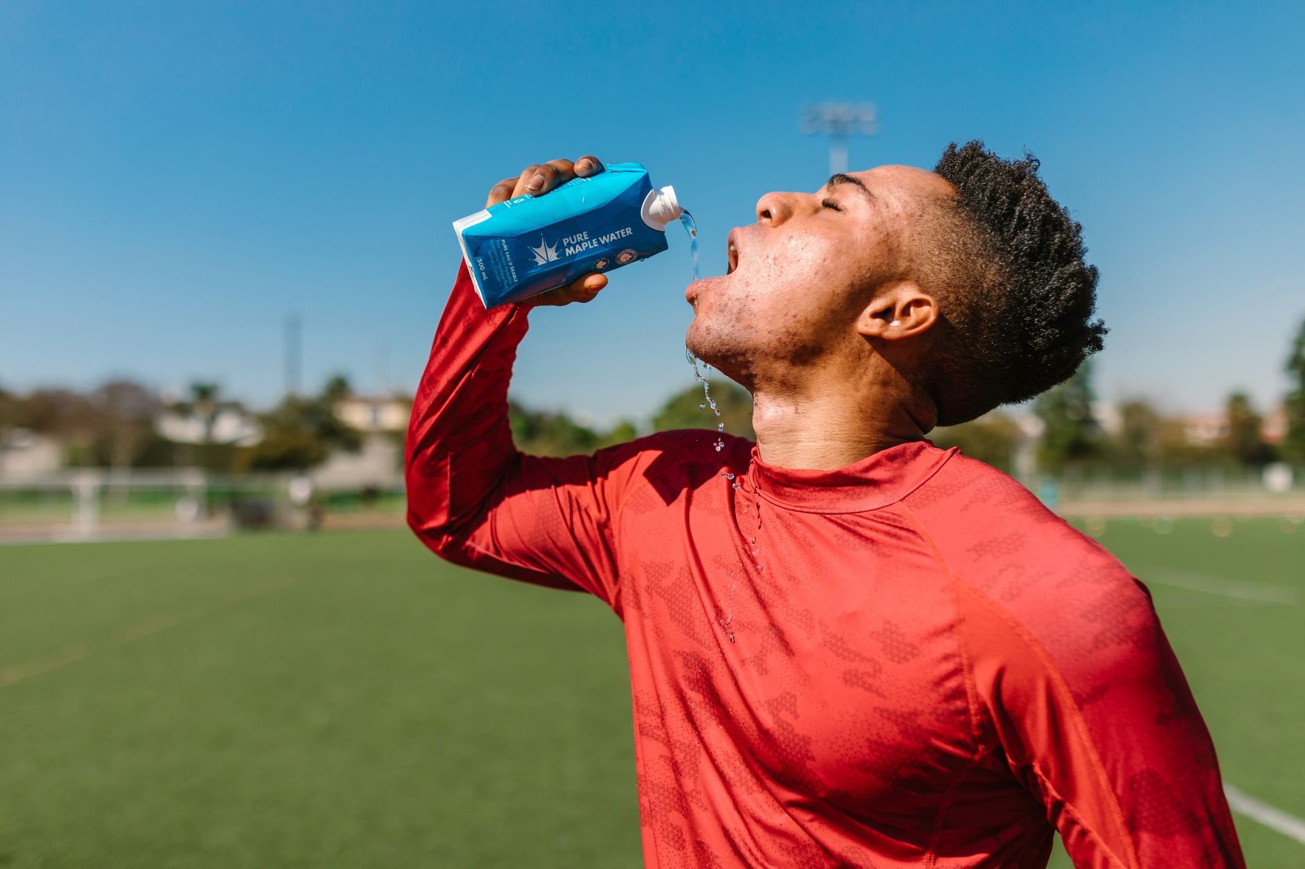 Athlet in rotem Sportshirt trinkt Wasser auf einem Sportplatz unter blauem Himmel.