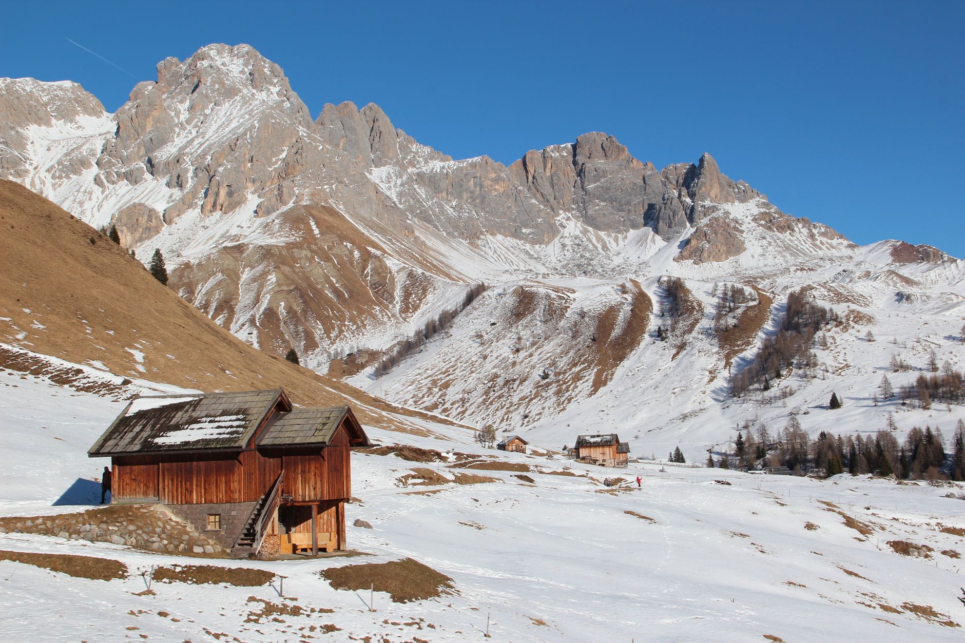 Red wooden chalet in snowy mountain valley with jagged peaks and larch forests under clear blue sky