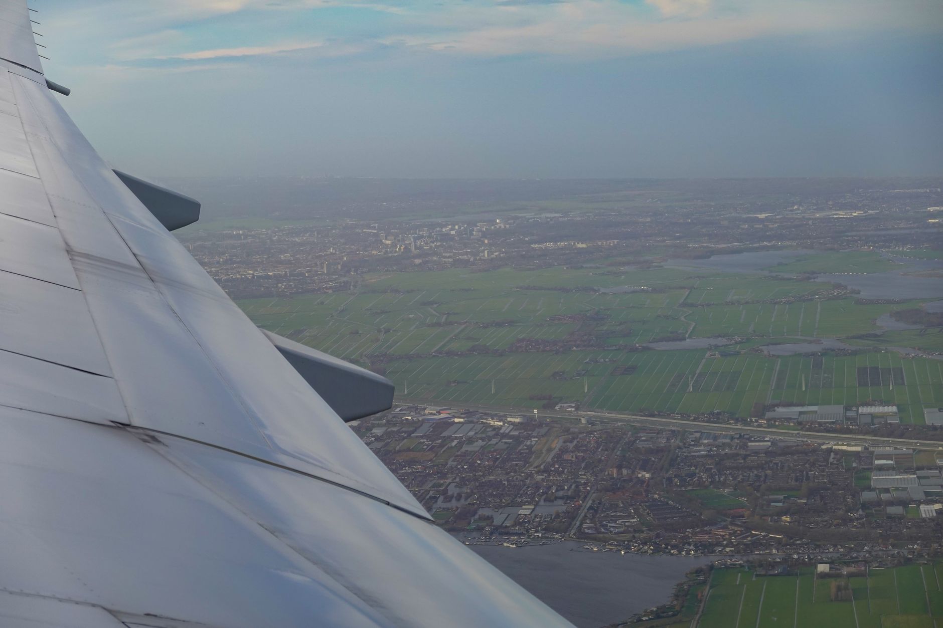 Aerial view from aircraft window showing wing and patchwork of green agricultural fields with urban development below