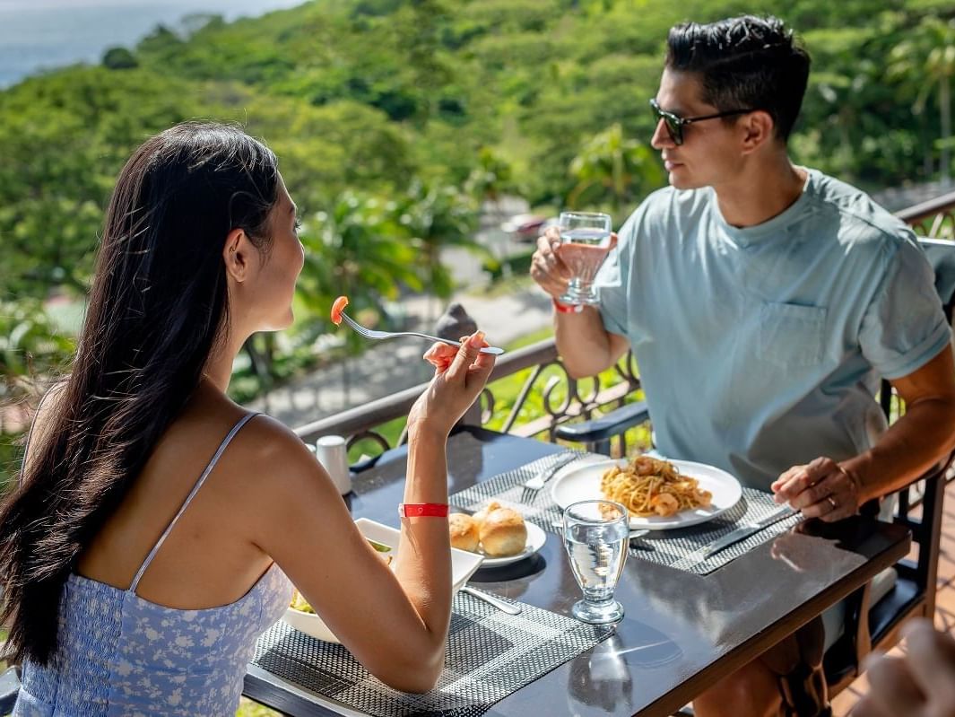 Couple dining on elevated terrace with plates of pasta and bread, enjoying scenic valley and ocean views with tropical vegetation