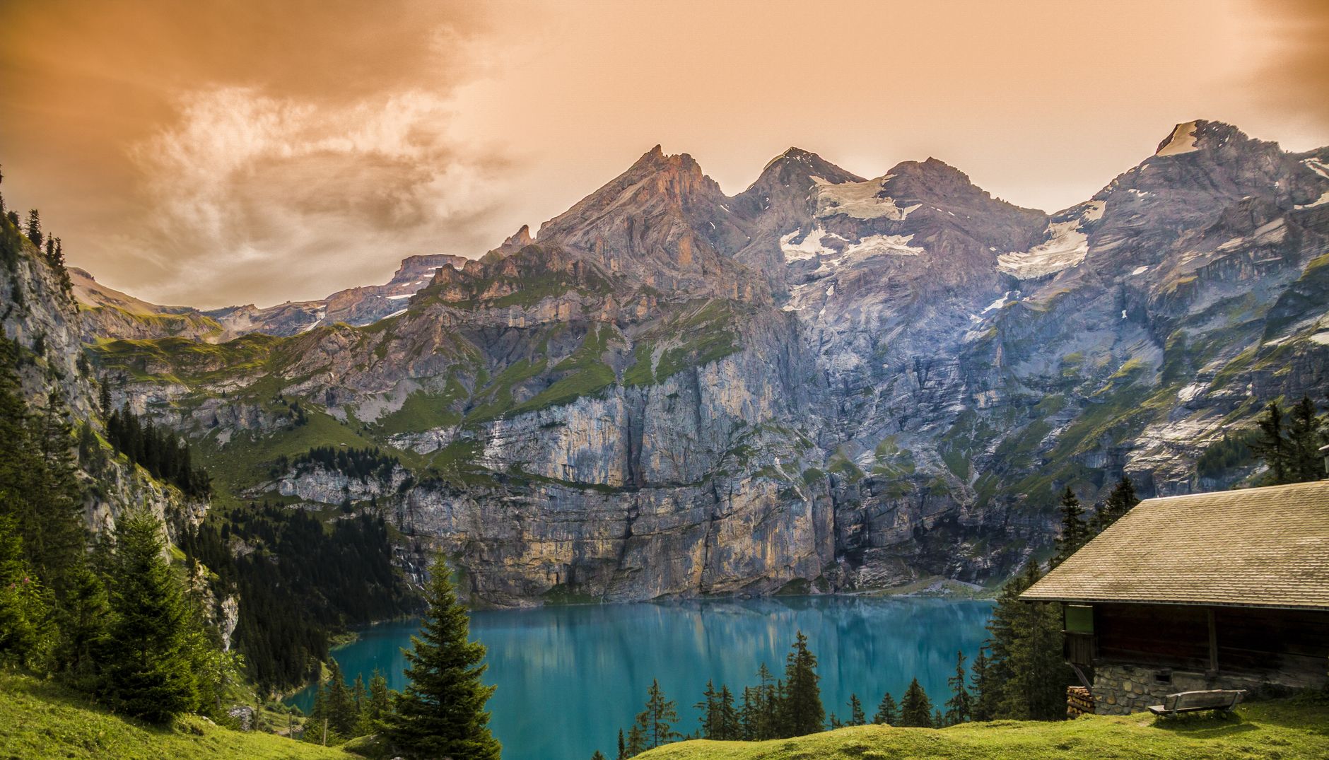 Turquoise alpine lake surrounded by dramatic rocky peaks, forested slopes, and a wooden cabin under golden sky