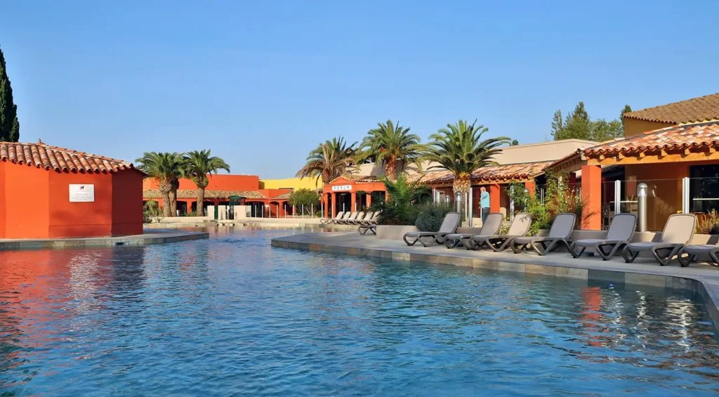 Piscine avec reflet de bâtiment orange, palmiers et transats gris au soleil.