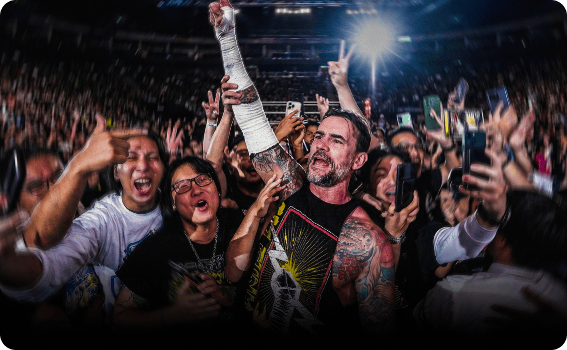 Fans celebrating in a stadium crowd, with one person holding up a cast-wrapped arm, surrounded by cheering supporters and bright stadium lights