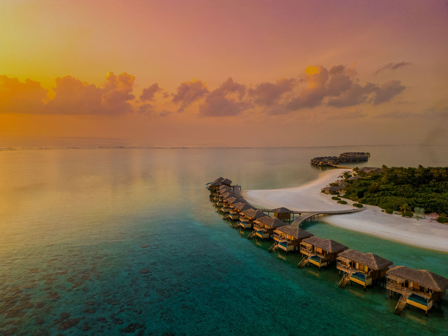 Overwater bungalows at sunset with turquoise water, white sand beach, and tropical island vegetation under golden and pink sky