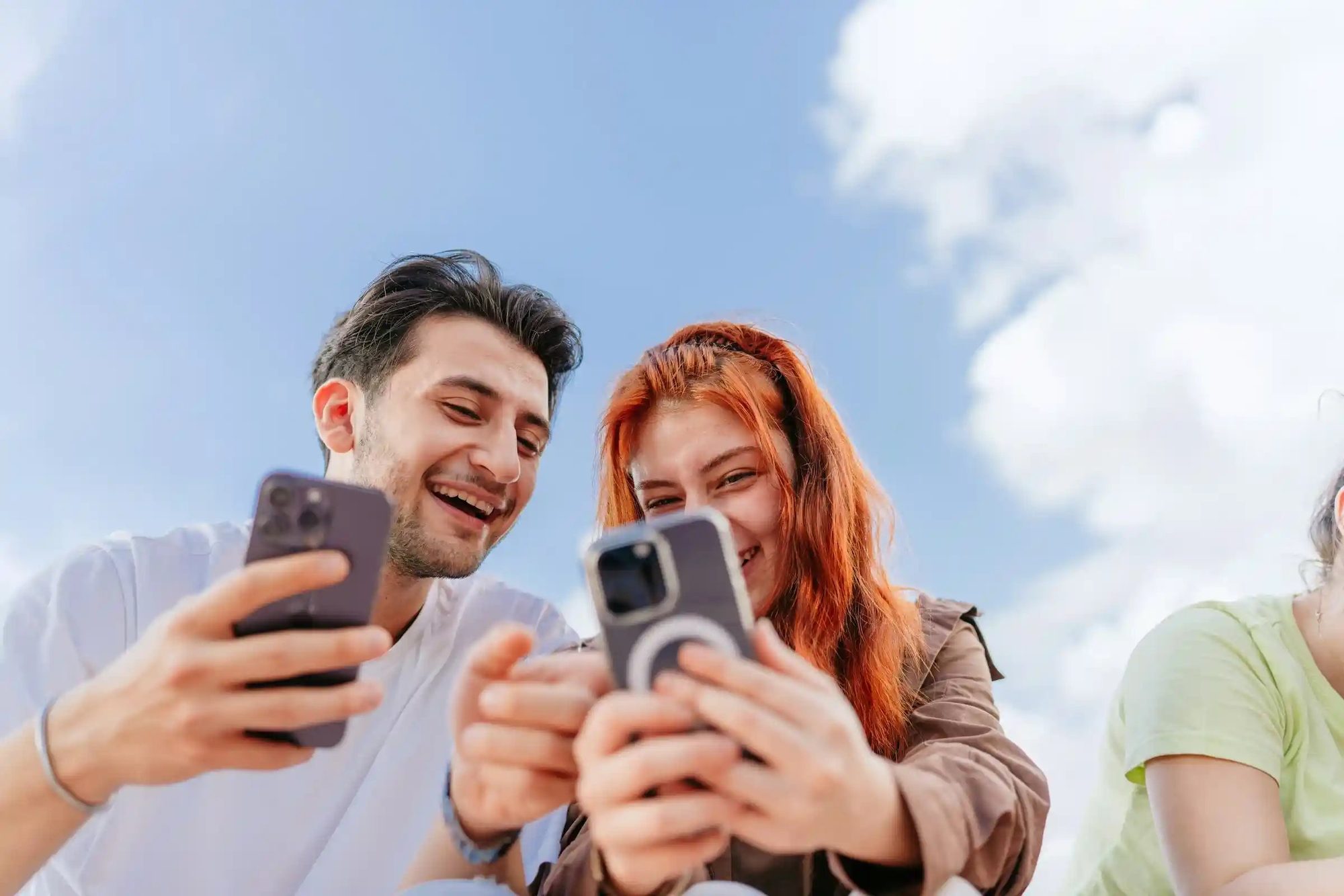 Friends outdoors holding smartphones and laughing together under blue sky with clouds