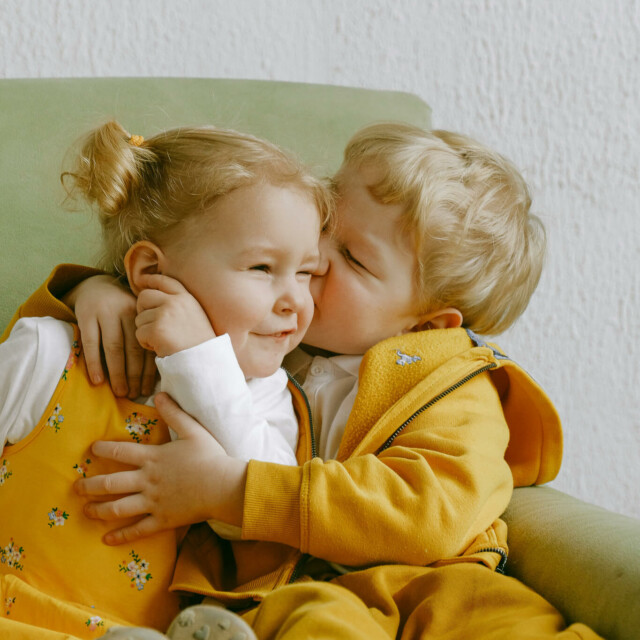 Two young blonde children in yellow outfits hugging on green chair