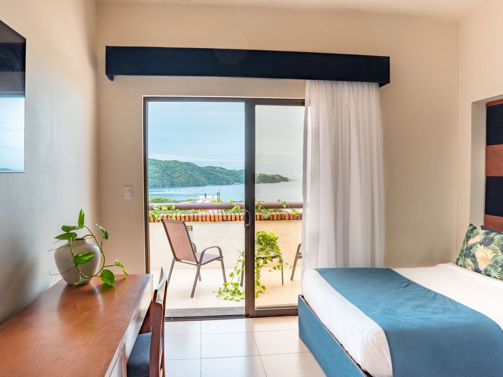 Beachfront villa bedroom with blue-accented bed, wooden desk, sliding glass doors opening to ocean view and patio with lounge chairs