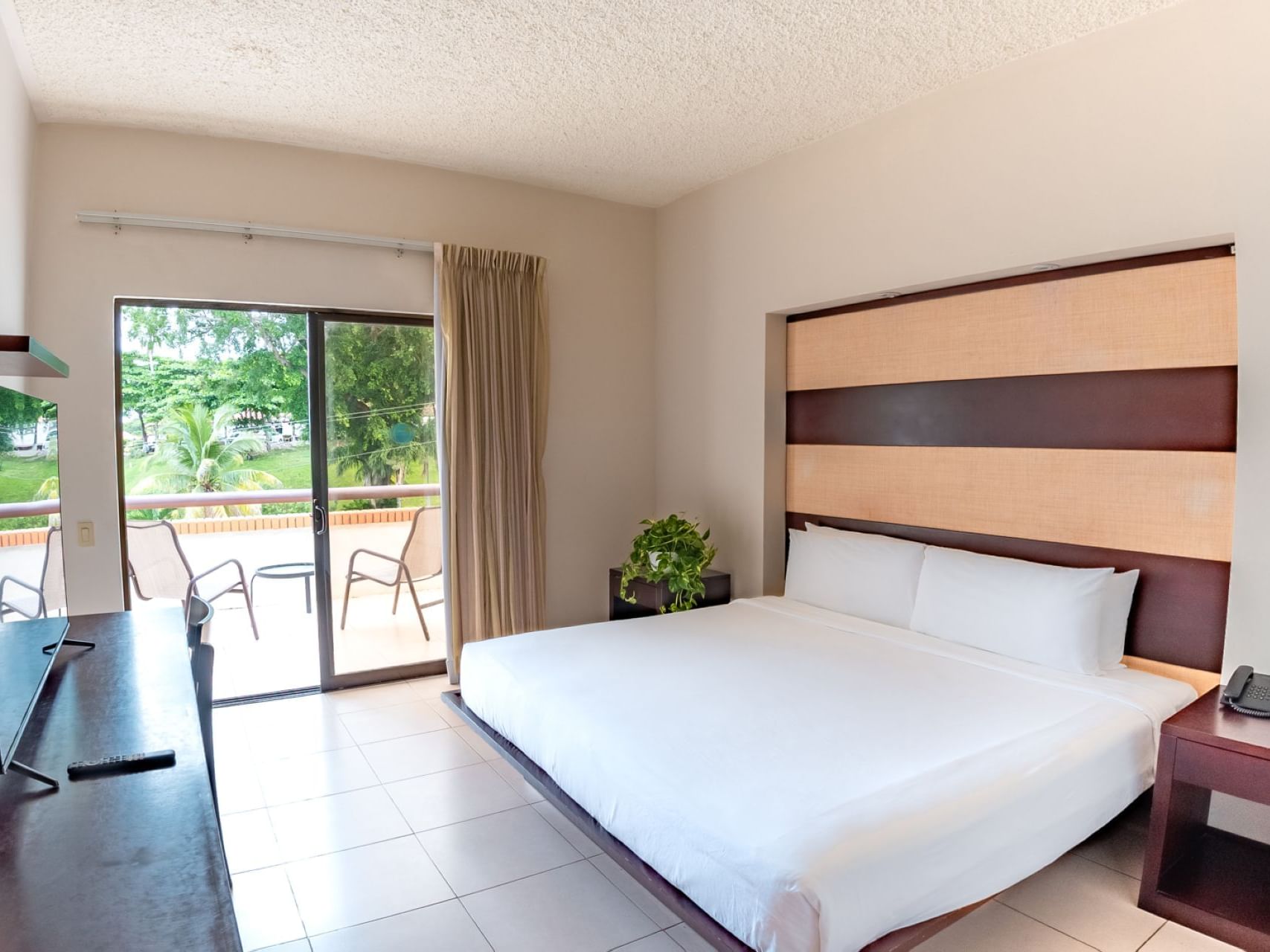 Spacious hotel room with striped wooden headboard, white bedding, glass door to balcony with lush greenery and distant water view