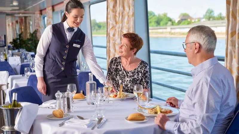 Couple dînant au restaurant d'un bateau de croisière avec vue sur l'eau et serveur en uniforme