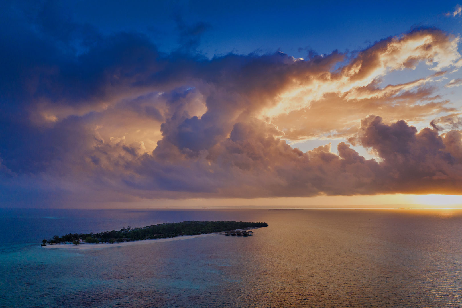 Aerial view of small island at sunset with pristine white beach, dense tropical vegetation, and dramatic golden clouds over calm sea