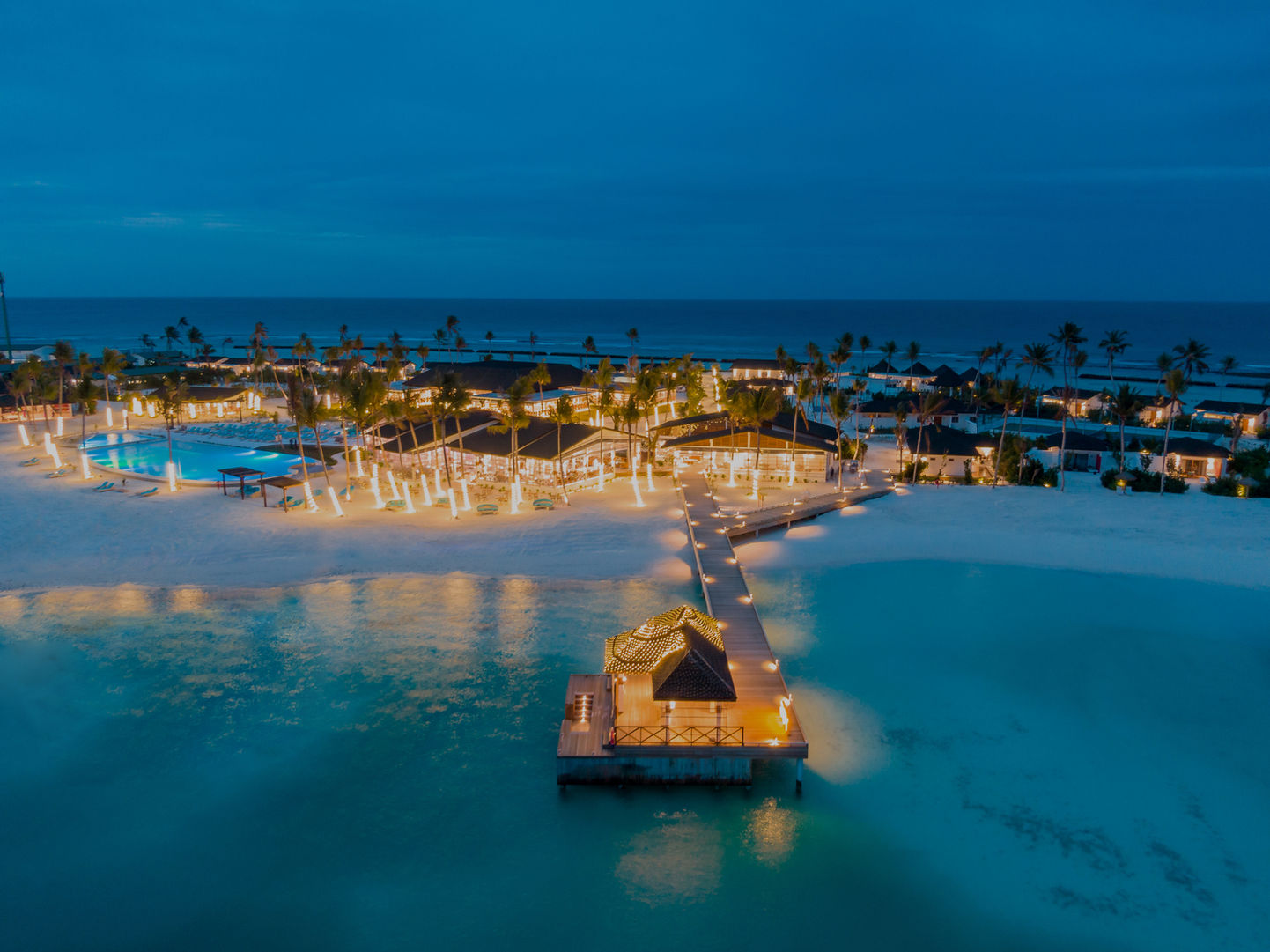 Illuminated resort pool complex at dusk with palm trees, white sand areas, and calm turquoise ocean under deep blue sky
