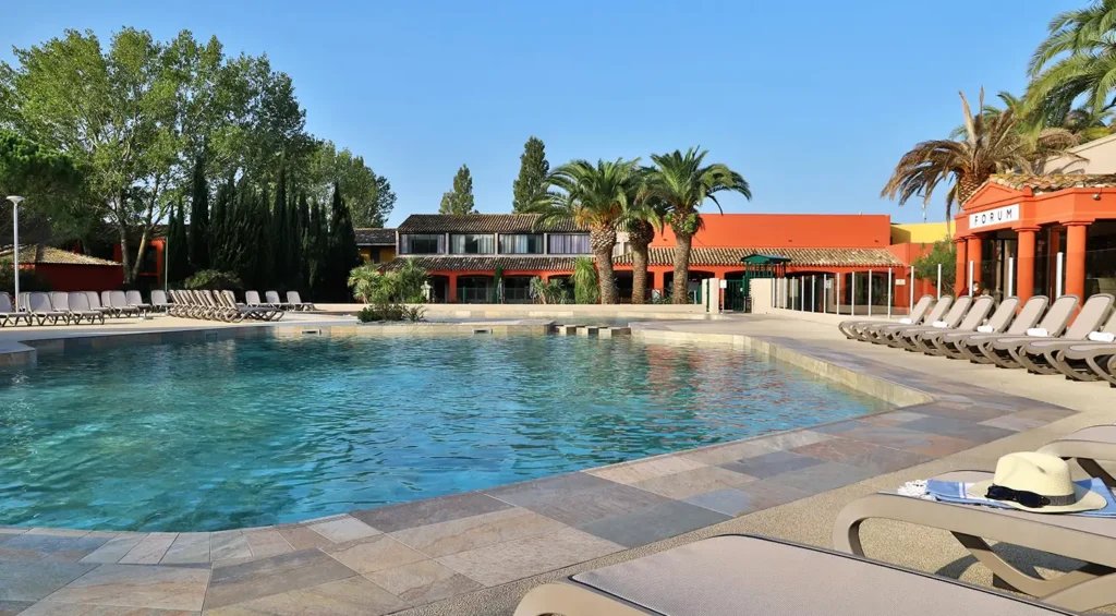Piscine rectangulaire avec bâtiments oranges, palmiers et chaises longues beiges.