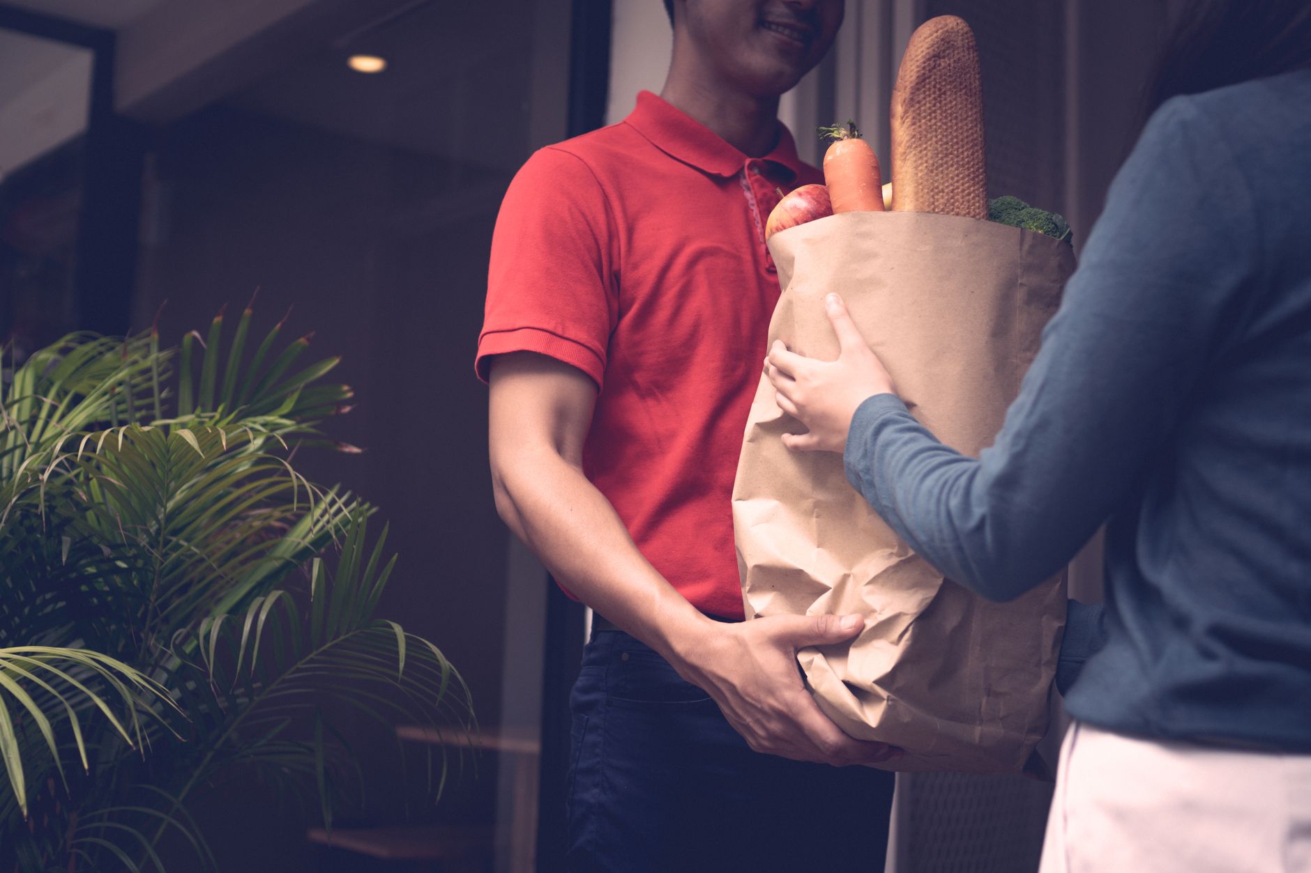 Livreur en chemise rouge tenant un sac en papier kraft rempli de provisions à la porte d'un client