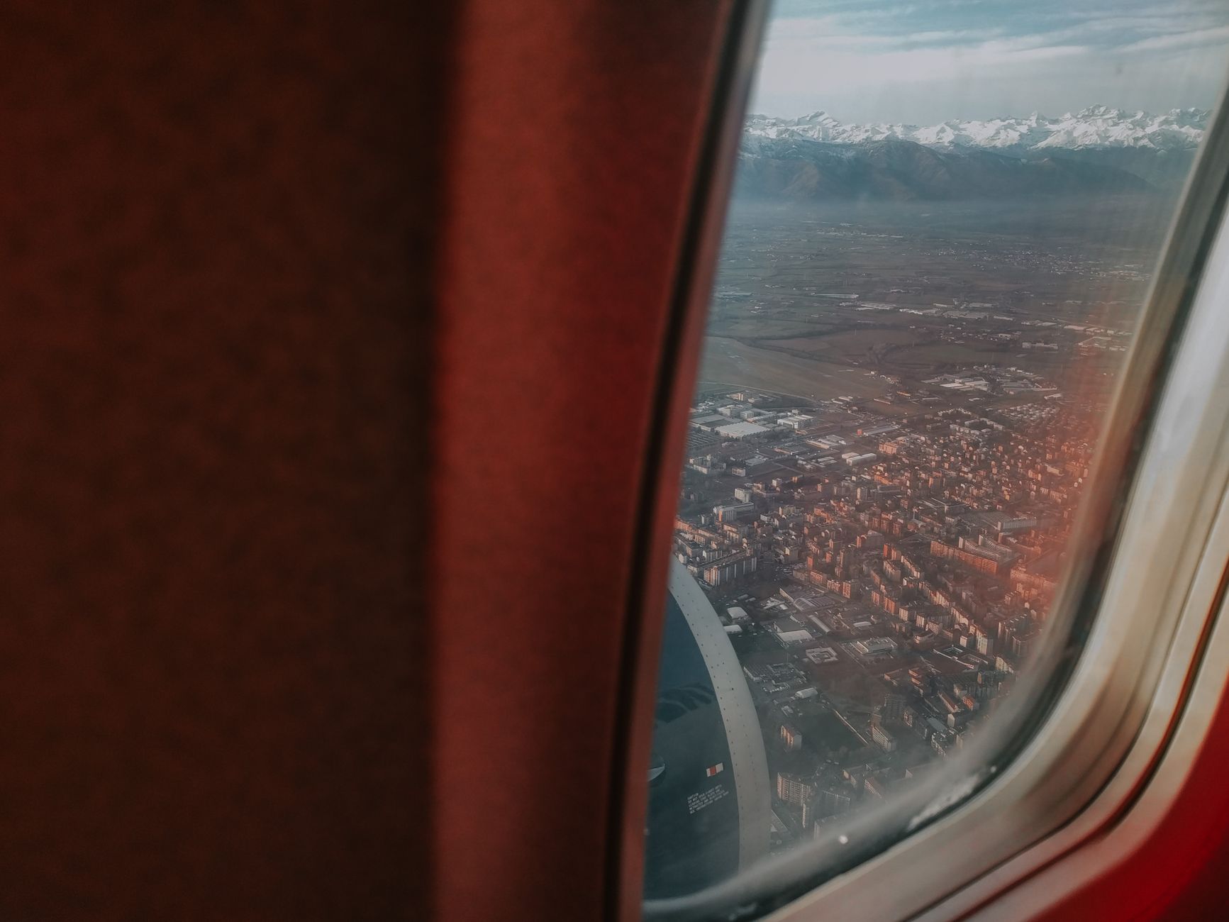 Airplane window view with red interior fabric showing snow-capped mountains in distance and urban sprawl with buildings below