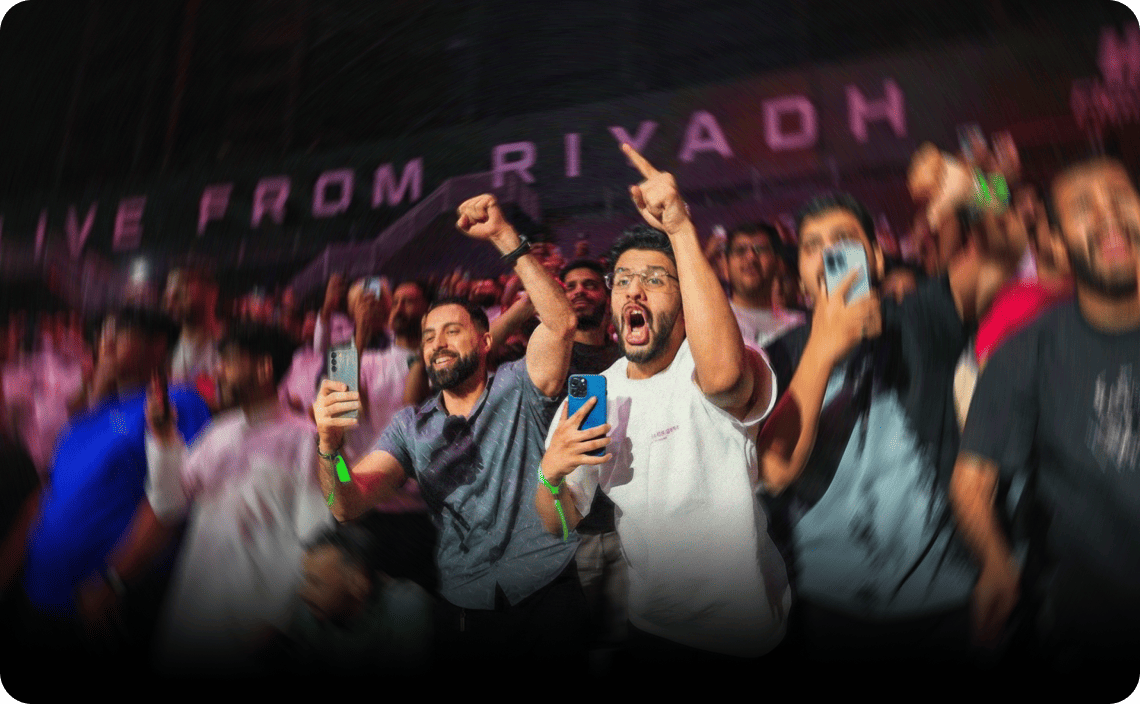 Crowd of enthusiastic spectators with hands raised, some holding phones, at an indoor venue with purple stage lighting and signage